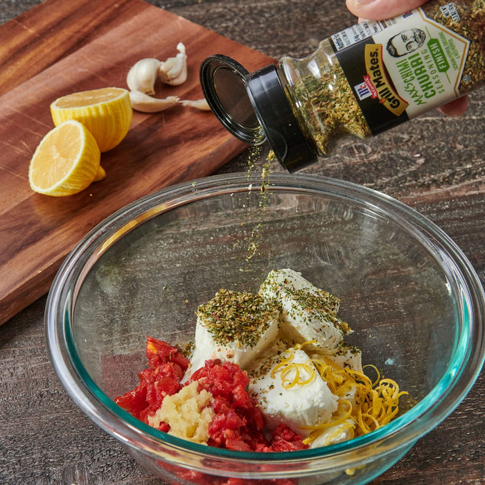 ['A glass bowl with ingredients for a dip, including tomatoes, garlic, cheese, and lemon zest, with a McCormick Grill Mates seasoning bottle pouring seasoning on top.']