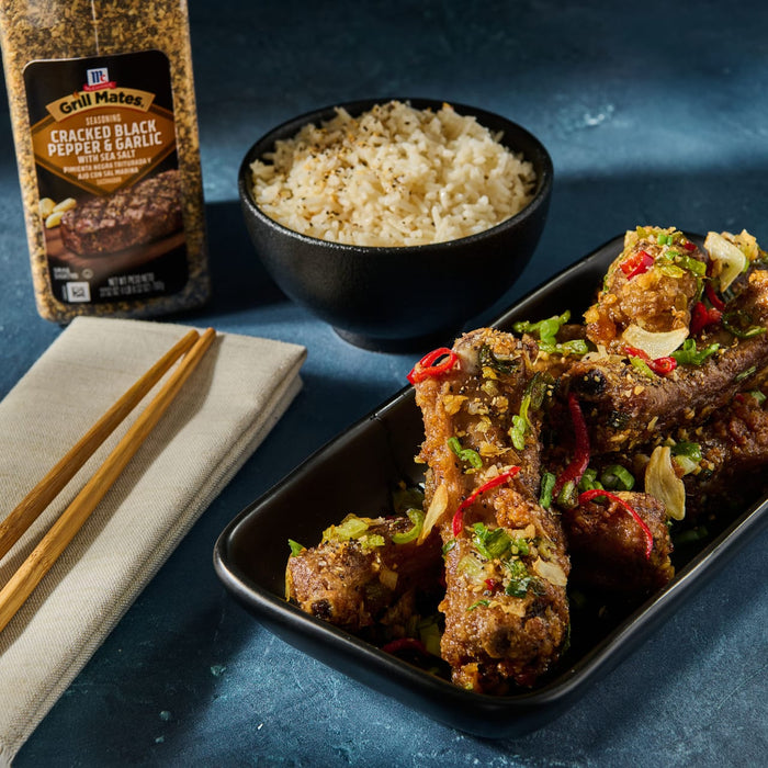 ['A plate of crispy pork ribs with garlic, black pepper, and chili peppers, a bowl of rice, and a bottle of Grill Mates seasoning.']