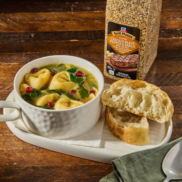 ['A bowl of soup with tortellini, spinach, and pomegranate seeds, a plate with two pieces of bread, a spoon, and a container of McCormick Grill Mates Cracked Black Pepper &amp; Garlic seasoning.']