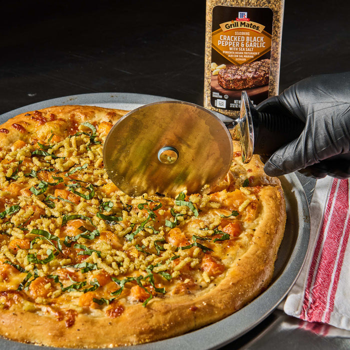 ['A pizza topped with butternut squash, basil, and a sprinkle of McCormick Grill Mates Cracked Black Pepper &amp; Garlic seasoning is being sliced with a pizza cutter.']