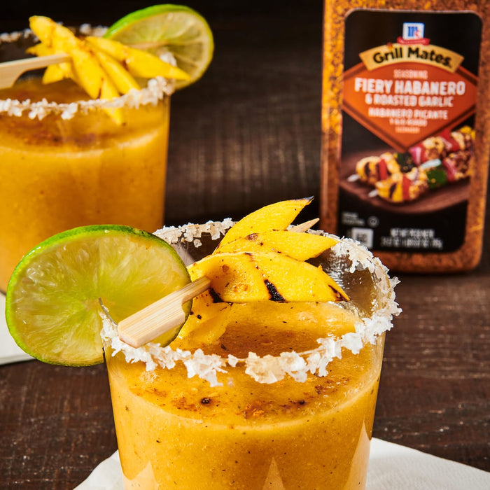 ['A glass of mango margarita with a lime wedge and grilled mango slices, next to a bottle of Grill Mates Fiery Habanero &amp; Roasted Garlic seasoning.']