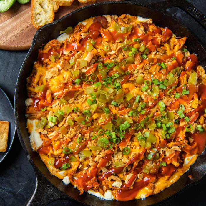 A skillet filled with a cheesy dip topped with shredded chicken, hot sauce, and green onions, served with toasted bread and sliced cucumbers.