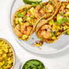 Three shrimp tacos with pineapple salsa and cilantro are arranged on a white plate, with a bowl of pineapple salsa and a bowl of cilantro nearby.