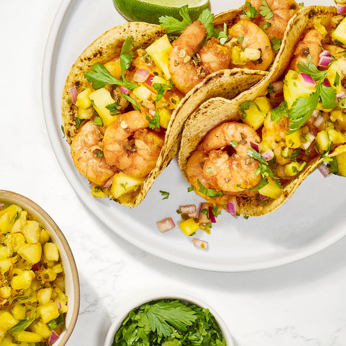 Three shrimp tacos with pineapple salsa and cilantro are arranged on a white plate, with a bowl of pineapple salsa and a bowl of cilantro nearby.