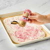 A hand sprinkles Great Value brand red sprinkles onto a white frosted dessert in a baking pan.
