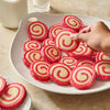 A hand reaches for a pinwheel cookie from a plate filled with many more, alongside glasses of milk.