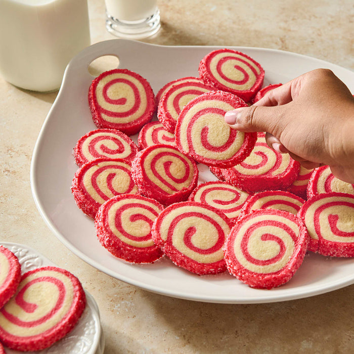 A hand reaches for a pinwheel cookie from a plate filled with many more, alongside glasses of milk.