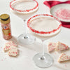 Two festive cocktails with red-sprinkled rims are displayed alongside pieces of white chocolate bark and a McCormick brand sprinkles container.