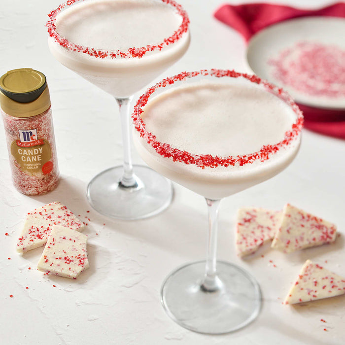 Two festive cocktails with red-sprinkled rims are displayed alongside pieces of white chocolate bark and a McCormick brand sprinkles container.