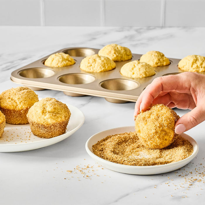 A hand dips a cornbread muffin into a plate of cinnamon sugar, with a muffin tin and a plate of muffins in the background.