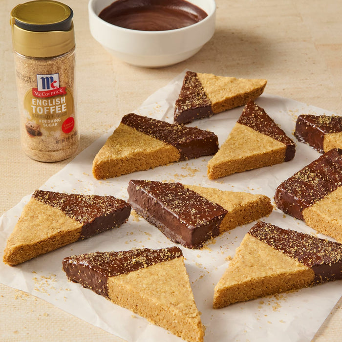 A batch of triangle-shaped shortbread cookies, half dipped in chocolate and sprinkled with McCormick brand cinnamon sugar, are arranged on parchment paper next to a bowl of chocolate sauce and a McCormick cinnamon sugar container.