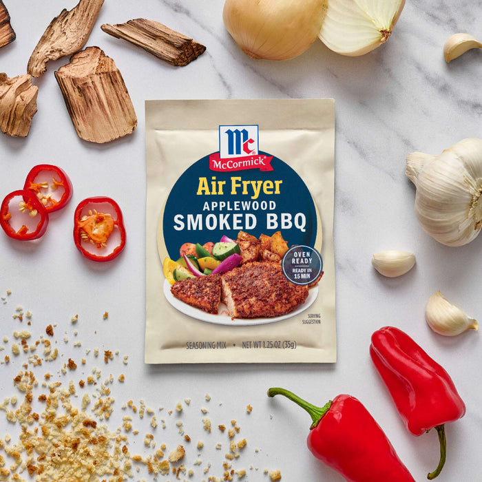 A packet of McCormick Air Fryer Applewood Smoked BBQ seasoning is displayed amidst ingredients like peppers, garlic, and wood chips.