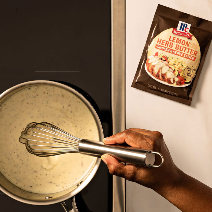 A person whisks a creamy sauce in a saucepan, prepared using the Mccormick Lemon Herb Butter All Purpose Sauce Mix Envelope .87 Oz packet shown on the counter.