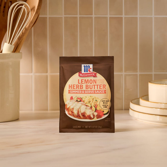 A single packet of Mccormick Lemon Herb Butter All Purpose Sauce Mix Envelope .87 Oz is displayed on a kitchen counter, with kitchen utensils and plates visible in the background to suggest a home-cooking setting.