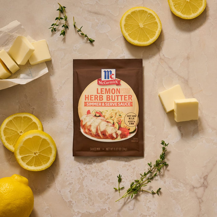 A flat lay image shows a packet of Mccormick Lemon Herb Butter All Purpose Sauce Mix Envelope .87 Oz surrounded by fresh ingredients like sliced lemons, pats of butter, and sprigs of thyme on a marble surface.