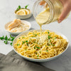 A creamy sauce made from a Mccormick Lemon Herb Butter All Purpose Sauce Mix Envelope .87 Oz is being poured from a glass jar over a bowl of spaghetti garnished with fresh parsley.