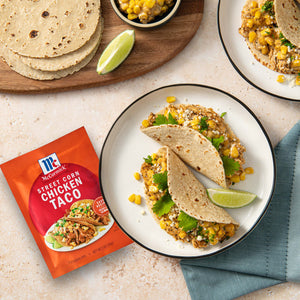 A packet of McCormick Perfectly Seasoned Chicken Taco seasoning is displayed next to plates of chicken tacos with corn and lime wedges.