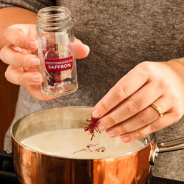 A person's hands are shown, one holding a jar of Mccormick Gourmet Saffron 0.035 Oz and the other dropping saffron threads into a steaming copper pot of liquid.