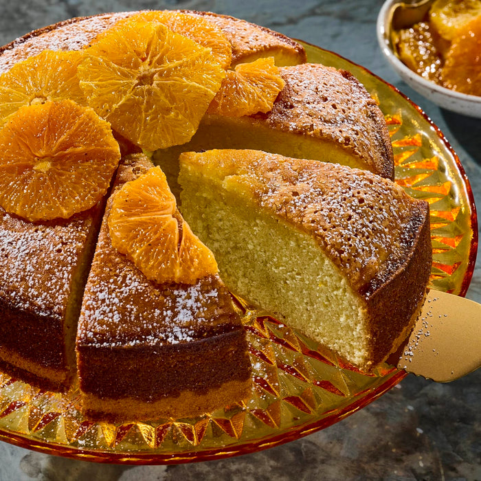 A round orange cake, dusted with powdered sugar and adorned with fresh orange slices, has a slice being served from a golden plate, representing a delightful dessert that could be prepared with the exquisite Mccormick Gourmet Saffron 0.035 Oz for a unique flavor.
