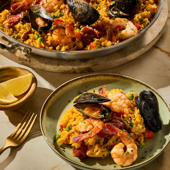 A serving of paella with shrimp, mussels, and sausage is plated next to a pan of the same dish, accompanied by lemon wedges.