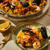 A serving of paella with shrimp, mussels, and sausage is plated next to a pan of the same dish, accompanied by lemon wedges.