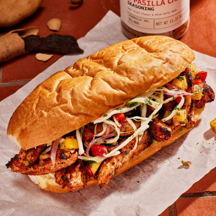 ['A close-up of a sandwich with grilled chicken, pineapple, red onion, and cabbage, with a jar of Pasilla Chile Seasoning in the background.']