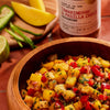 ['A wooden bowl filled with a mango salsa with a jar of Tamarind &amp; Pasilla Chili Seasoning in the background.']