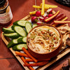 ['A bowl of hummus with various toppings, surrounded by cucumber, carrot, radish, and pita bread.']