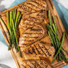 ['Grilled chicken breasts and asparagus spears on a wooden cutting board.']