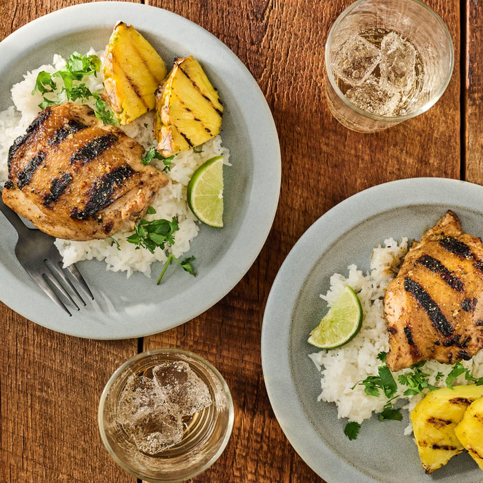 Overhead shot of two plates of grilled chicken with rice pineapple and lime accompanied by glasses of a clear beverage with ice on a wooden table.