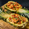 Two halves of a pineapple filled with shrimp and rice are displayed on a wooden board.