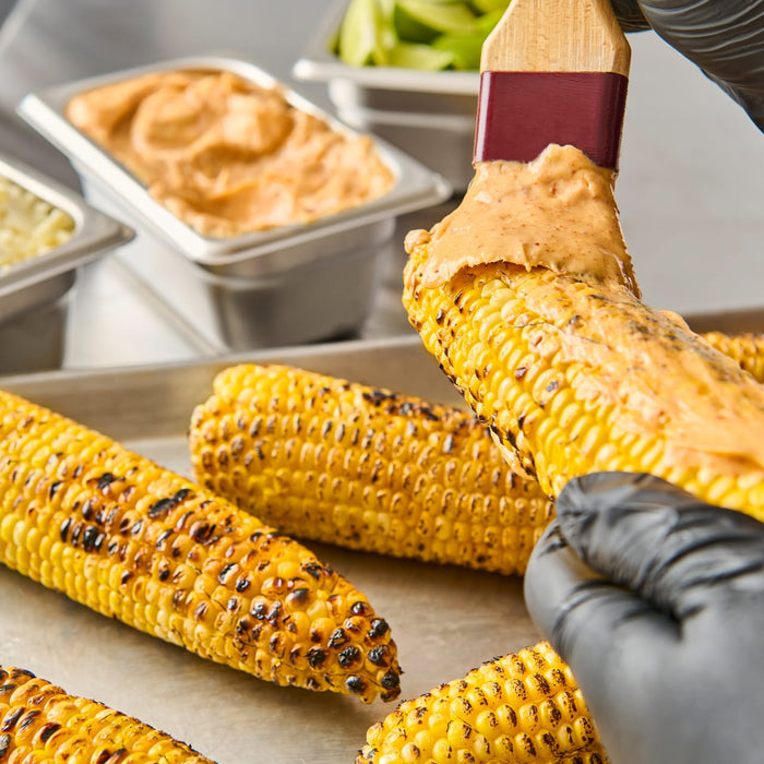 A gloved hand spreads a creamy sauce over a grilled corn cob.