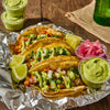 ['A close-up of four tacos topped with cilantro, onions, and a green sauce, served on a bed of aluminum foil.']
