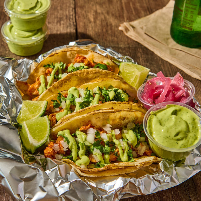 ['A close-up of four tacos topped with cilantro, onions, and a green sauce, served on a bed of aluminum foil.']