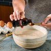 A person adds Nielsen-Massey vanilla extract to a bowl of batter while preparing a recipe.