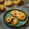 A plate of freshly baked chocolate chip cookies, some broken open to show the melted chocolate, with more cookies cooling on a wire rack in the background.
