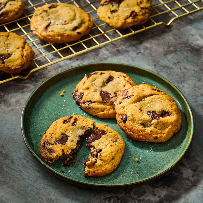 A plate of freshly baked chocolate chip cookies, some broken open to show the melted chocolate, with more cookies cooling on a wire rack in the background.