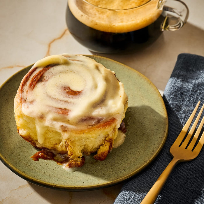 A frosted cinnamon roll sits on a plate next to a cup of coffee, suggesting a delicious breakfast or treat.