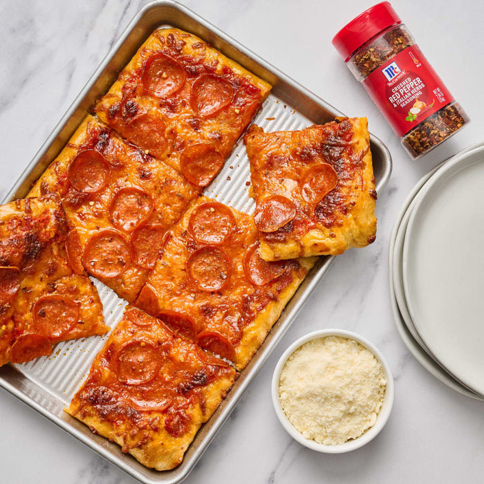 ['A baking sheet with pepperoni pizza slices, a small bowl of grated parmesan cheese, and a jar of McCormick Crushed Red Pepper &amp; Italian Herbs Seasoning.']
