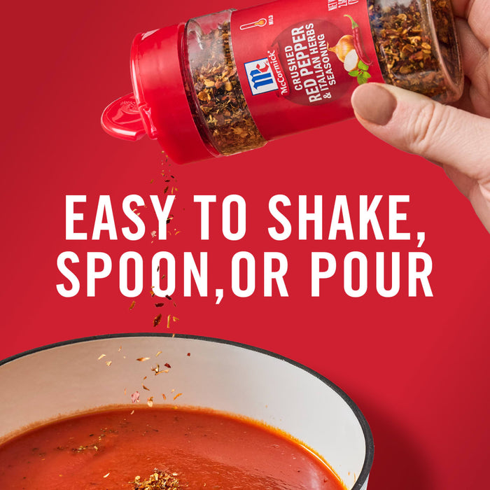 ['A hand holds a McCormick Crushed Red Pepper &amp; Italian Herbs Seasoning bottle over a bowl of tomato soup.']