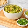 ['A bowl of guacamole with tortilla chips, lime wedges, and cilantro on a wooden cutting board.']