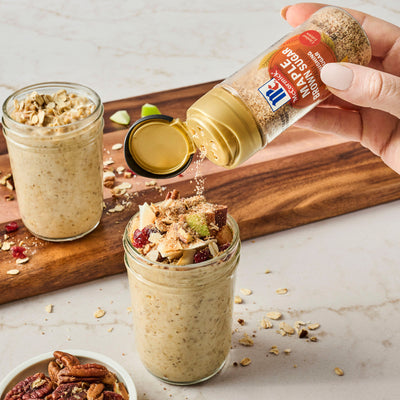 Hand pouring McCormick Maple Brown Sugar Seasoning into a jar of overnight oats with a wooden board and ingredients in the background.