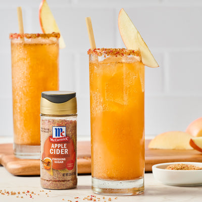 Two glasses of orange juice with apple slices, a bottle of McCormick Apple Cider Finishing Sugar, and apples on a wooden board.