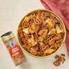 A bowl of apple-cinnamon snack mix with pretzels, pecans, and cereal pieces is displayed next to a bottle of McCormick Apple Pie Spice.