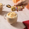 Two mugs of pumpkin spice lattes are being sprinkled with McCormick Pumpkin Pie Spice.