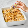 A hand sprinkles McCormick cinnamon sugar blend over a bread pudding in a white baking dish.