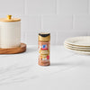 A bottle of McCormick Pumpkin Spice seasoning sits on a white countertop next to a decorative jar and stack of plates.