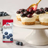 A box of McCormick Blueberry Extract is displayed next to mini cheesecakes topped with blueberries being drizzled with a blueberry sauce.