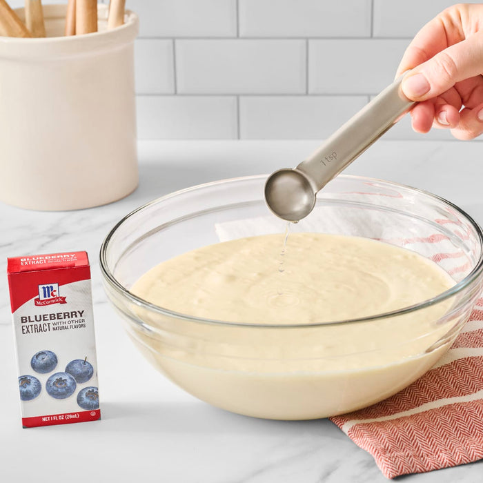 A hand adds liquid from a measuring spoon into a bowl of batter, next to a box of Mccormick Blueberry Extract 1 Fl Oz With Other Natural Flavors.