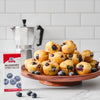 A box of Mccormick Blueberry Extract 1 Fl Oz With Other Natural Flavors stands on a white counter next to a wooden cake stand overflowing with blueberry muffins, with a silver moka pot in the background.